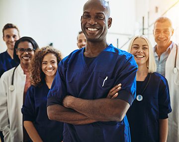 Young African male doctor smiling while standing in a hospital corridor with a diverse group of staff in the background