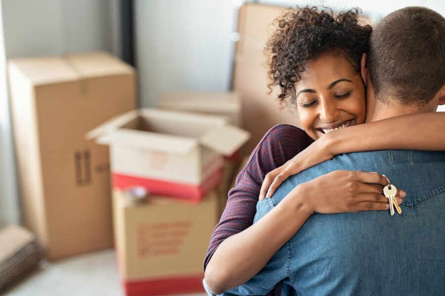 Woman with keys in her hand hugging a man with moving boxes in the background.