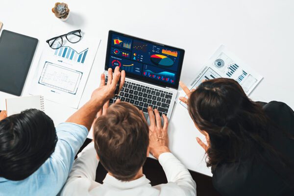 A photo of three individuals huddling around a laptop with charts and statistics.