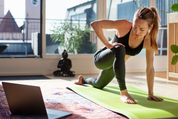 Image of a woman with her laptop and a yoga mat participating in a virtual yoga session.