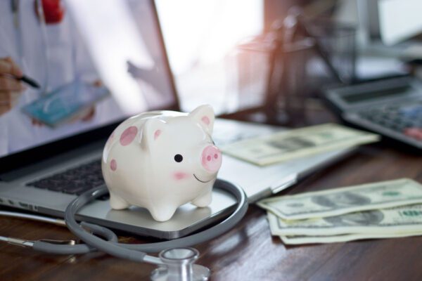 Photo of a piggy bank next to a doctor's stethoscope and a pile of cash.
